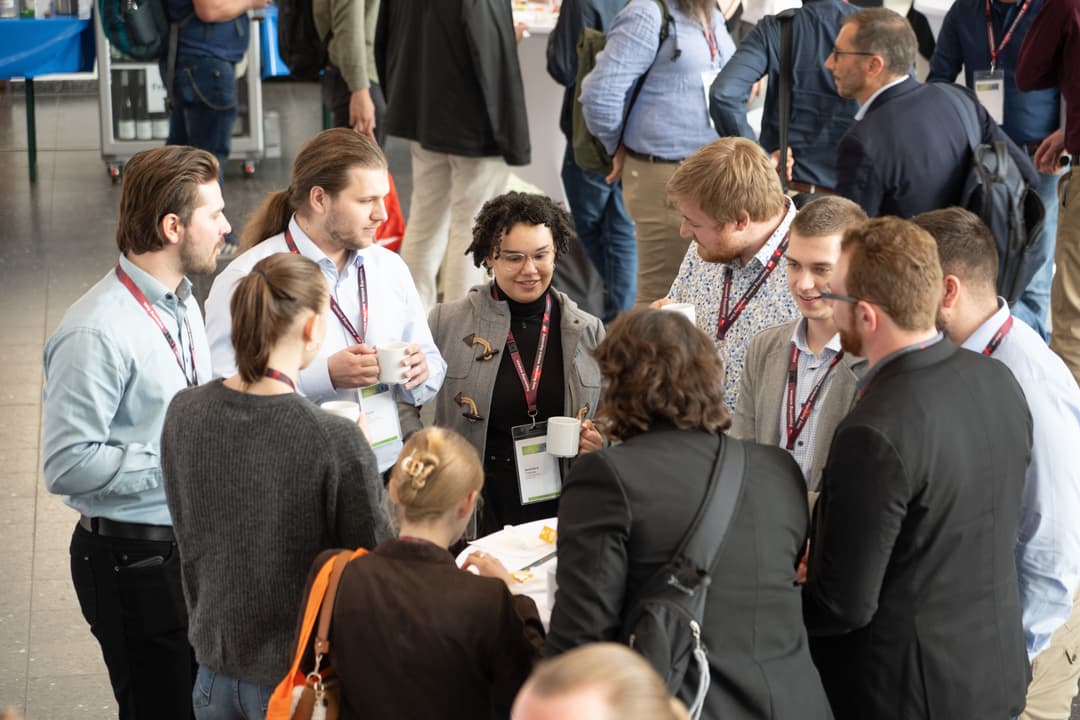 Gruppe von Konferenzteilnehmenden steht in der Kaffeepause in einem Foyer im Kreis, unterhält sich angeregt und hält Kaffeetassen in der Hand; alle tragen Namensschilder und Lanyards, im Hintergrund sind weitere Personen und Messestände zu sehen.