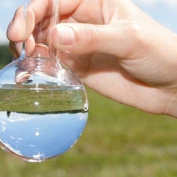 Hand hält rundkolbigen Glasbehälter mit klarer Flüssigkeit vor unscharf grünem Hintergrund