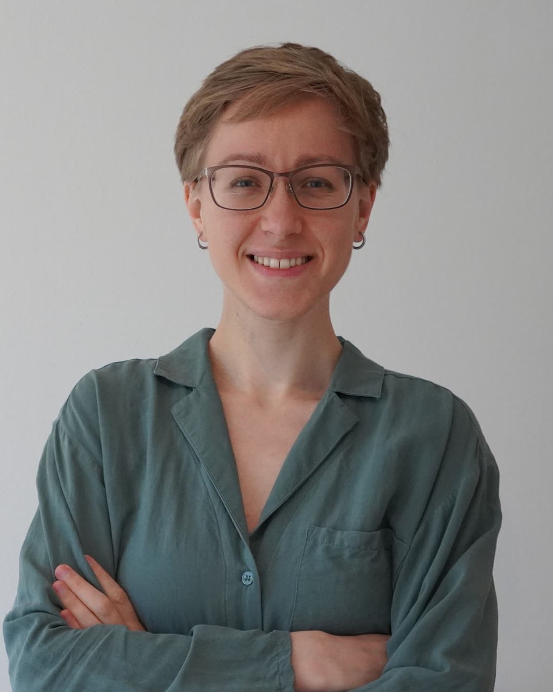 Alexandra Tietze lächelt freundlich in die Kamera. Sie trägt eine Brille und hat kurze blonde Haare. Ihre Arme sind vor der Brust verschränkt und sie trägt eine türkisfarbene Bluse mit Brusttasche. Der Hintergrund ist neutral grau-weiß gehalten. Das Porträt zeigt sie von der Brust aufwärts in professioneller Pose.