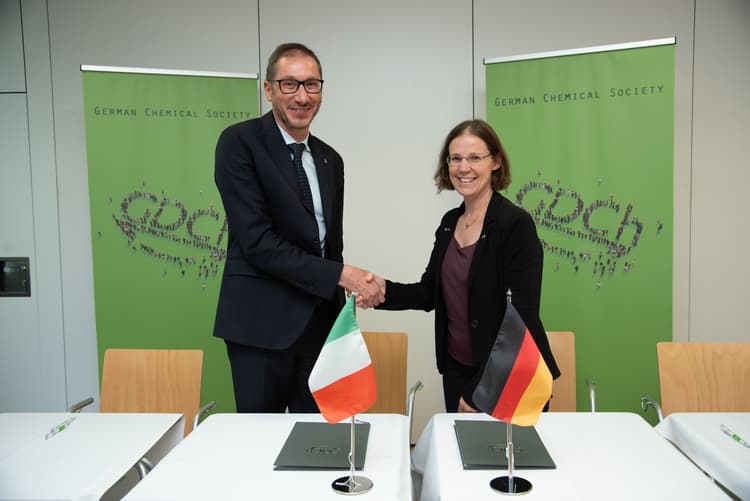 Zwei Personen stehen hinter einem Tisch und geben sich die Hand. Auf dem Tisch stehen zwei kleine Tischflaggen, eine mit den Farben Italiens und eine mit den Farben Deutschlands. Im Hintergrund sind zwei grüne Roll-ups mit dem Schriftzug 'German Chemical Society' und einem stilisierten Molekülmuster zu sehen. Die Szene spielt in einem Konferenzraum mit hellen Wänden und Holzstühlen.