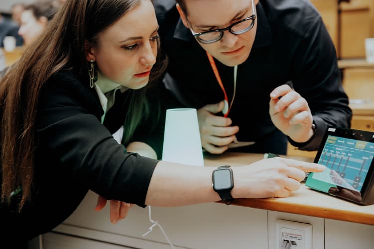 Eine Person mit langen braunen Haaren und schwarzer Kleidung zeigt mit dem Finger auf ein Tablet, das auf einer Tischplatte liegt. Das Tablet zeigt eine farbige Benutzeroberfläche mit mehreren Balkendiagrammen und Schaltflächen. Neben dem Tablet steht eine kleine, kegelförmige Lampe mit weißem Licht. Im Hintergrund sind weitere Personen und Möbelstücke zu erkennen.