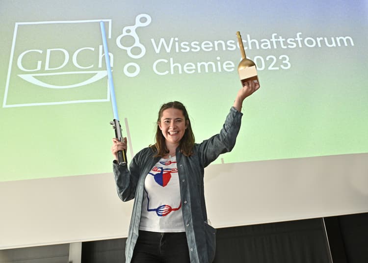 Eine Person steht vor einer großen Leinwand mit dem Schriftzug 'GDCh Wissenschaftsforum Chemie 2023'. Die Person hält in der rechten Hand eine goldene Trophäe und in der linken Hand mehrere lange Gegenstände. Sie trägt ein T-Shirt mit buntem Aufdruck und eine offene Jacke. Der Hintergrund zeigt ein Farbverlauf von Grün zu Blau.