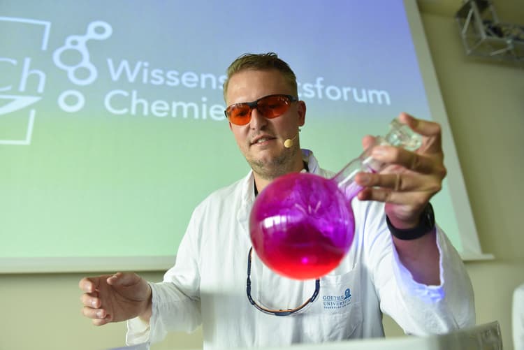Ein Chemiker im weißen Laborkittel und mit Schutzbrille präsentiert in einem Hörsaal einen runden Glaskolben mit leuchtend pinker Flüssigkeit; im Hintergrund ist auf der Leinwand das Logo und der Schriftzug „Wissenschaftsforum Chemie“ zu sehen.
