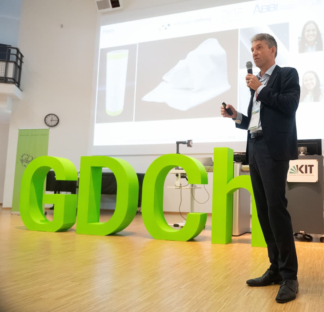 Buchstaben "GDCh" auf einer Bühne. Rechts eine Person bei der Präsentation eines wiss. Vortrags.