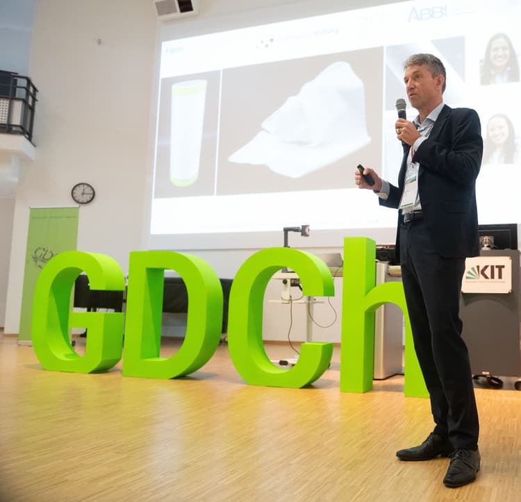 Buchstaben "GDCh" auf einer Bühne. Rechts eine Person bei der Präsentation eines wiss. Vortrags.