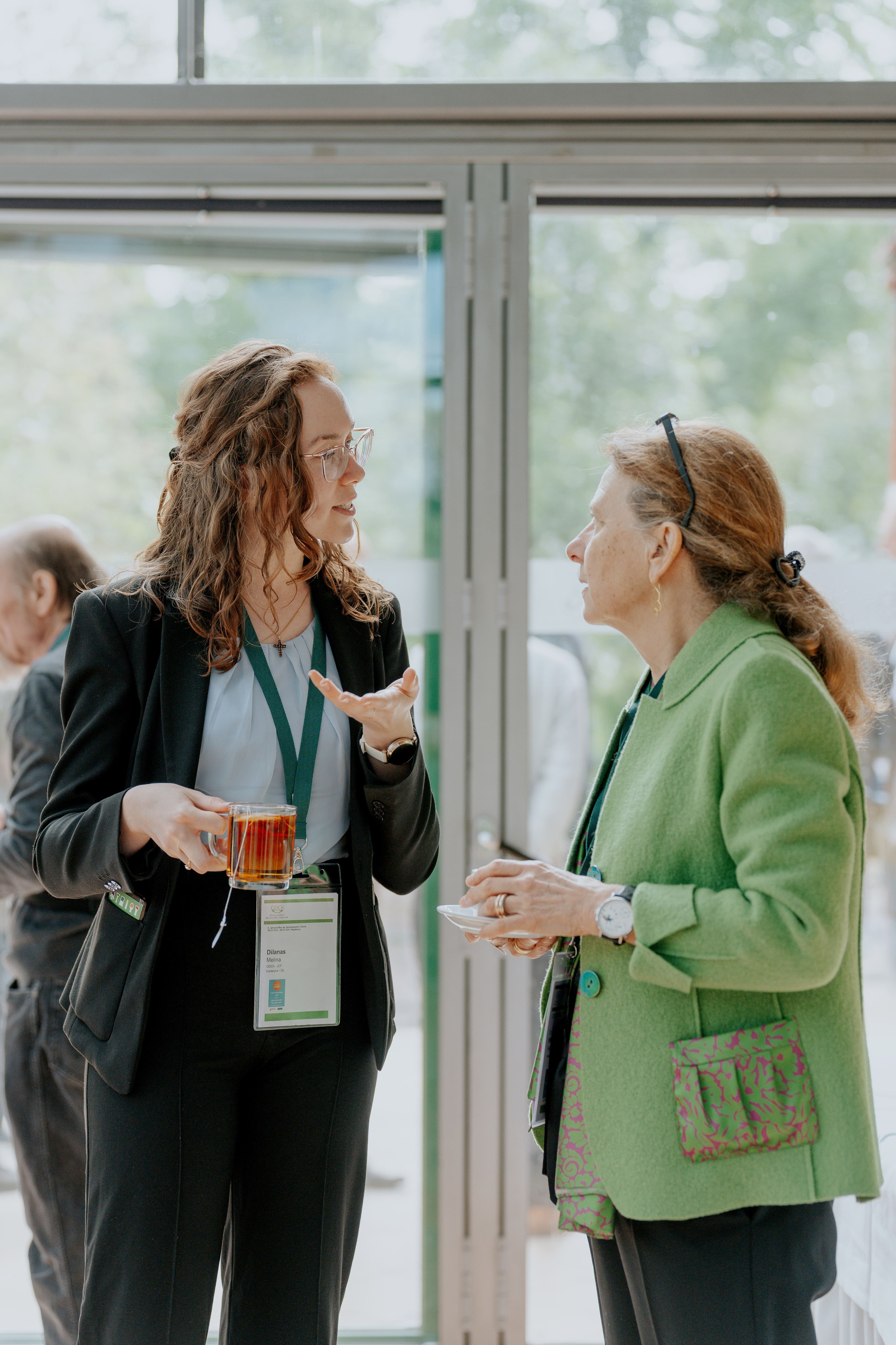 Zwei Personen stehen nebeneinander vor einer großen Glasfront. Die linke junge Person trägt einen schwarzen Hosenanzug und hält ein Glas mit einem roten Getränk in der rechten Hand. Die rechte ältere Person trägt eine grüne Jacke mit gemusterten Taschen und hält eine kleine weiße Tasse. Beide tragen Namensschilder an grünen Bändern um den Hals. Im Hintergrund sind weitere Personen und Bäume außerhalb des Gebäudes zu erkennen.