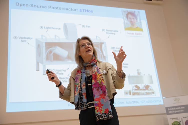 Frau mit buntem Schal hält Mikrofon und Präsentationsfernbedienung vor einer Projektionswand mit technischer Grafik und Text.