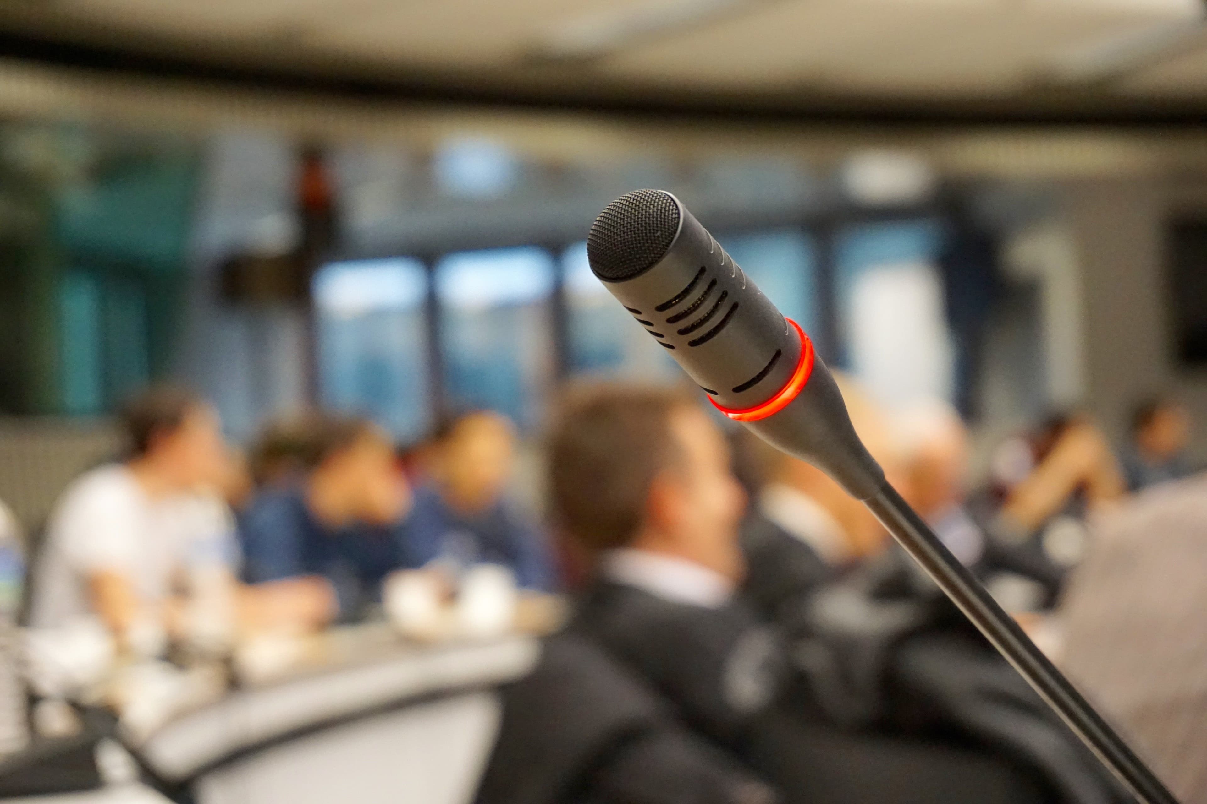 Nahaufnahme eines Mikrofonkopfs mit rotem Leuchtring vor unscharfem Hintergrund einer Veranstaltung mit mehreren Personen.