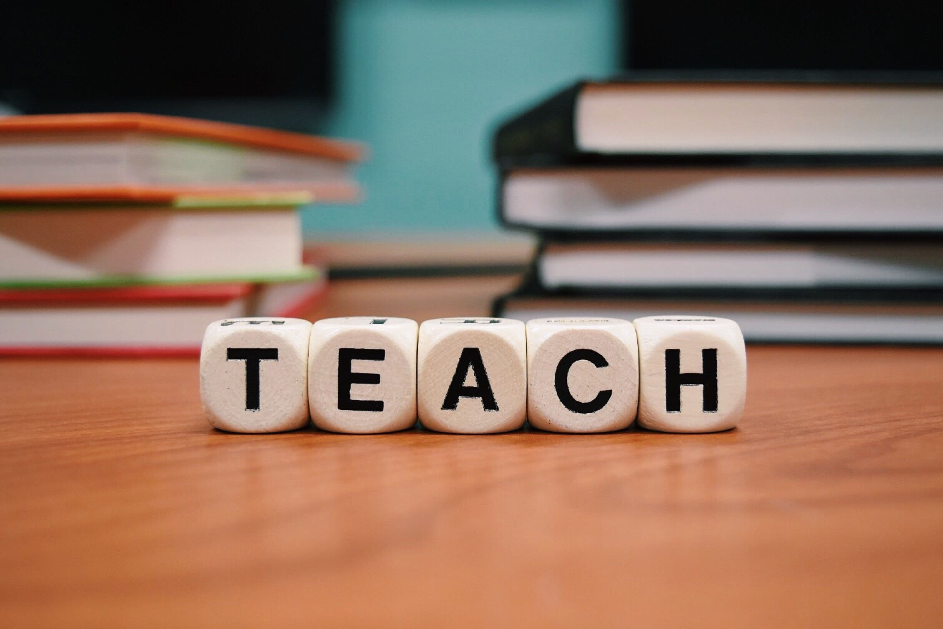 Würfel mit den Buchstaben T E A C H auf einem Holztisch vor unscharfem Hintergrund mit mehreren Bücherstapeln