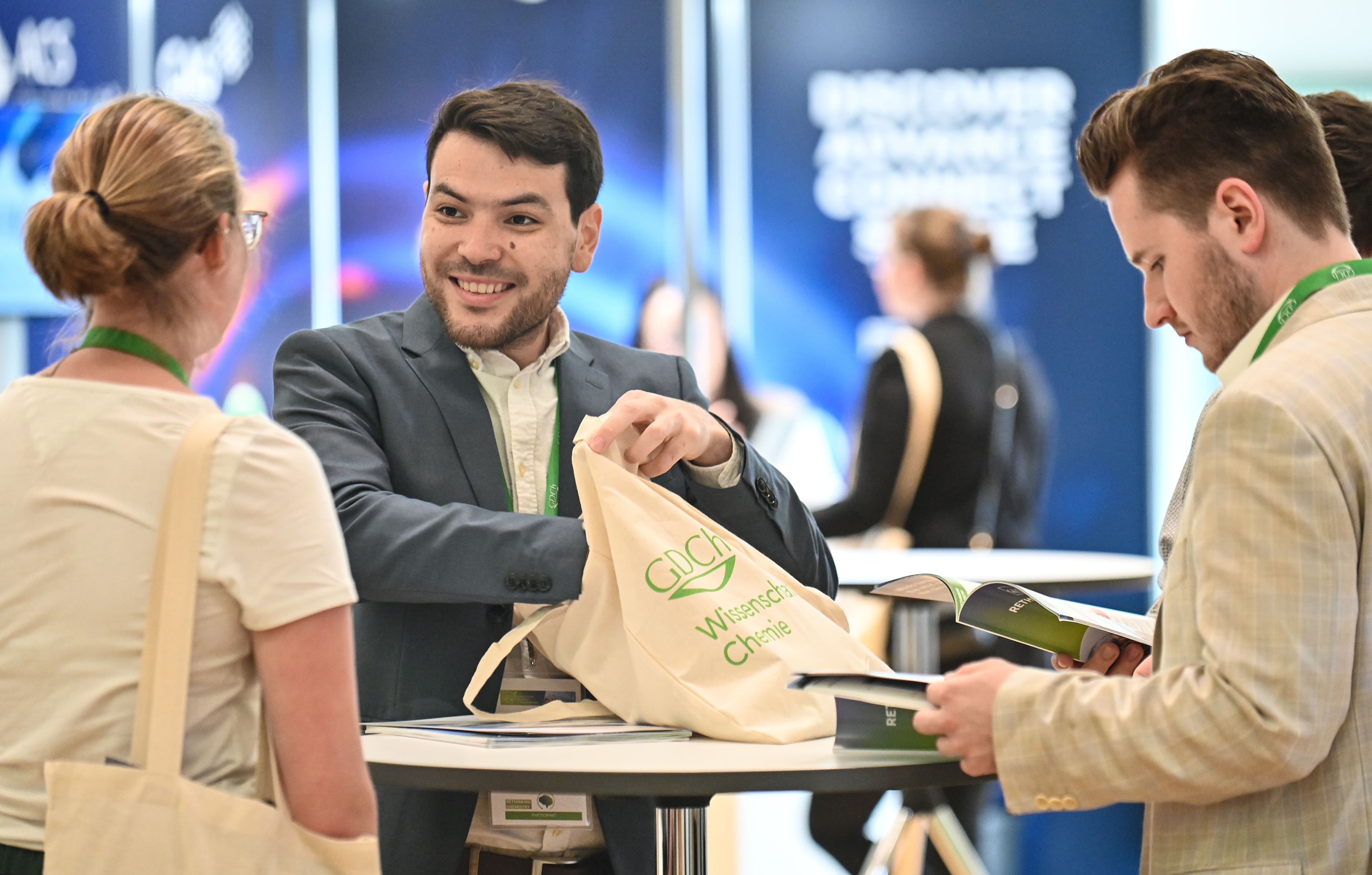Drei Personen stehen an einem runden Stehtisch in einer hellen Messe- oder Konferenzumgebung. Eine Person hält eine beigefarbene Stofftasche mit grünem Aufdruck, während die anderen beiden Broschüren oder Hefte in den Händen halten. Alle tragen grüne Namensschilder um den Hals. Im Hintergrund sind unscharfe Messestände mit blauen und weißen Flächen sowie weitere Personen zu sehen.