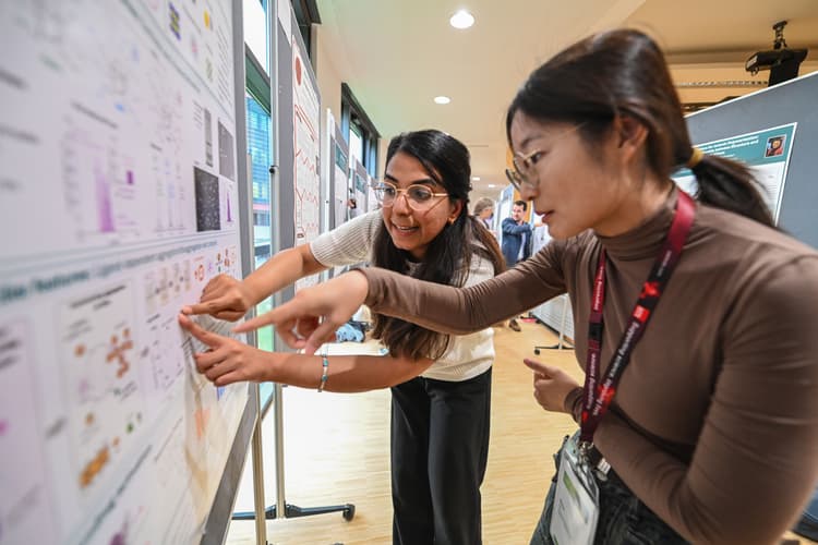 Zwei Teilnehmerinnen stehen vor einer Posterwand auf einer wissenschaftlichen Konferenz und zeigen auf Diagramme und Abbildungen, während sie die Inhalte diskutieren.