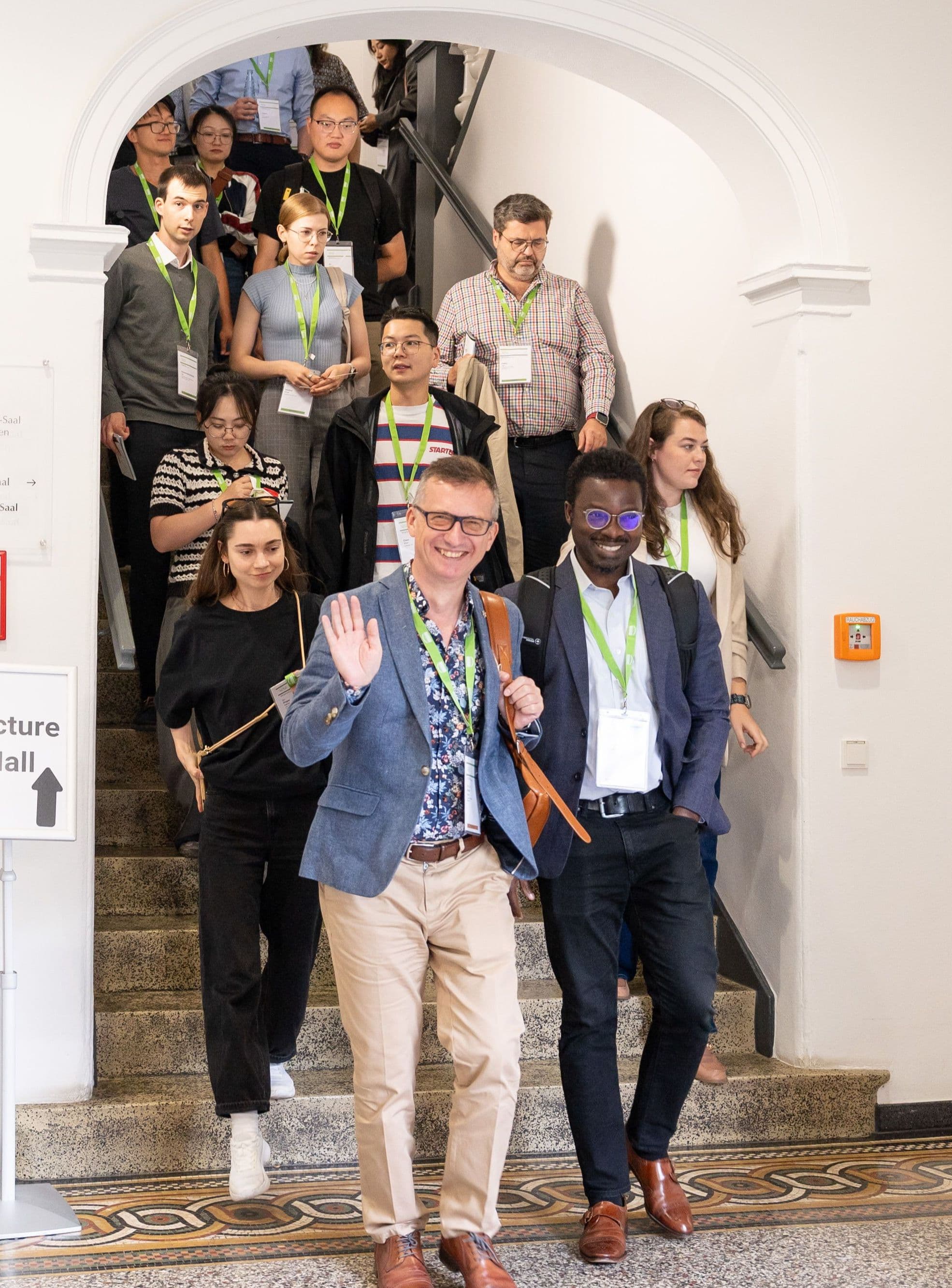 Eine Gruppe von Menschen mit Namensschildern um den Hals geht eine breite Steintreppe in einem hellen Gebäude hinunter. Im Vordergrund winkt eine Person und lächelt, während eine weitere Person daneben läuft. Die Menschen tragen überwiegend legere Kleidung. An der Wand links hängt ein Schild mit der Aufschrift 'Lecture Hall' und einem Richtungspfeil. Die Umgebung wirkt modern und gepflegt, mit weißen Wänden und Glastüren.